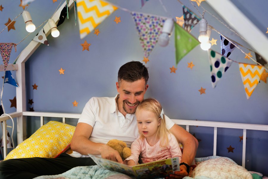 Young handsome father and his daughter reading a book while sitting in bed. Happy father reading book with his little girl while sitting on bed before bedtime together