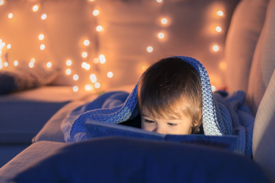 Cute little baby boy reading a book lying on sofa under knitted blanket with garland lights at background. Evening home leisure time. Christmas magic. Magical world of book.